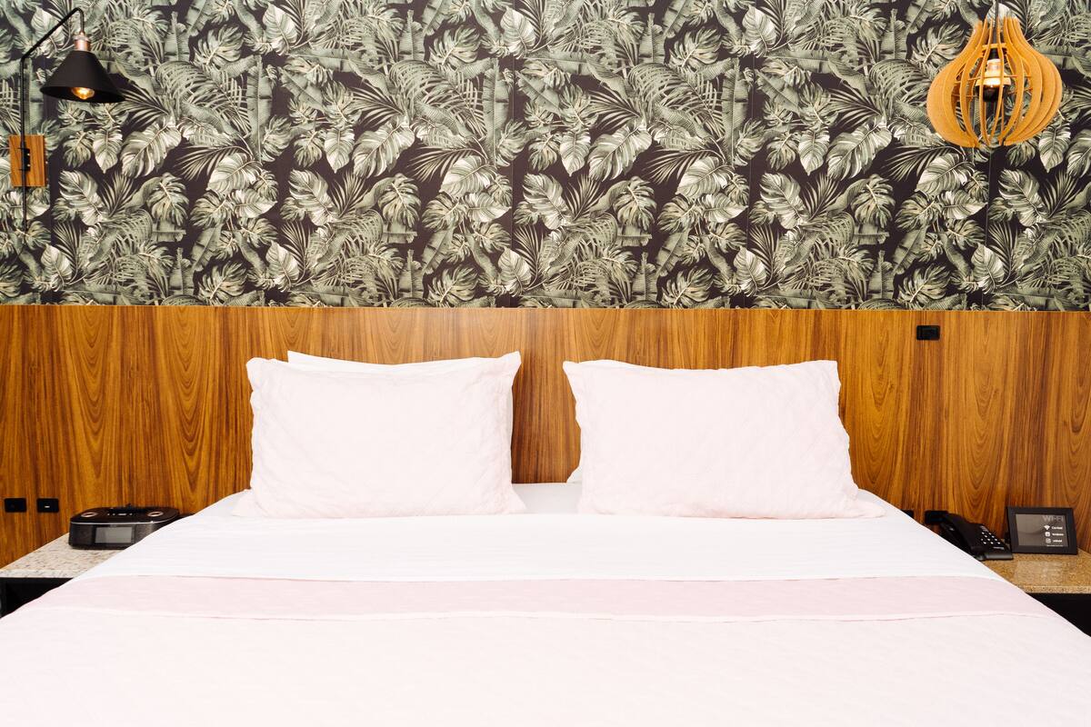 A hotel room with a wooden headboard, a bed with white linens, a patterned wallpaper, a black lamp, and a wooden hanging light fixture.