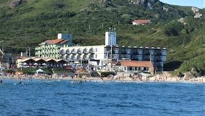 Un hotel sul lungomare con diversi piani, una spiaggia in primo piano e un paesaggio collinare sullo sfondo.