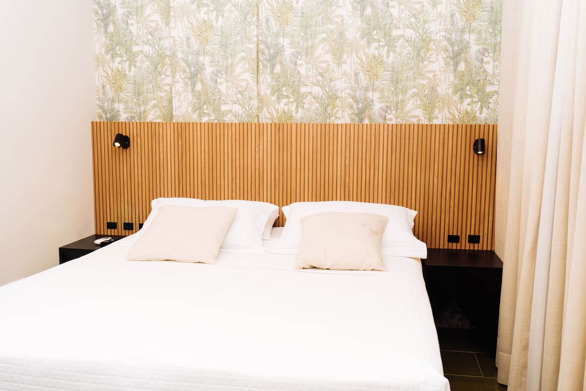 A hotel room with a wooden headboard, two white pillows, a white bedspread, and a patterned wallpaper.