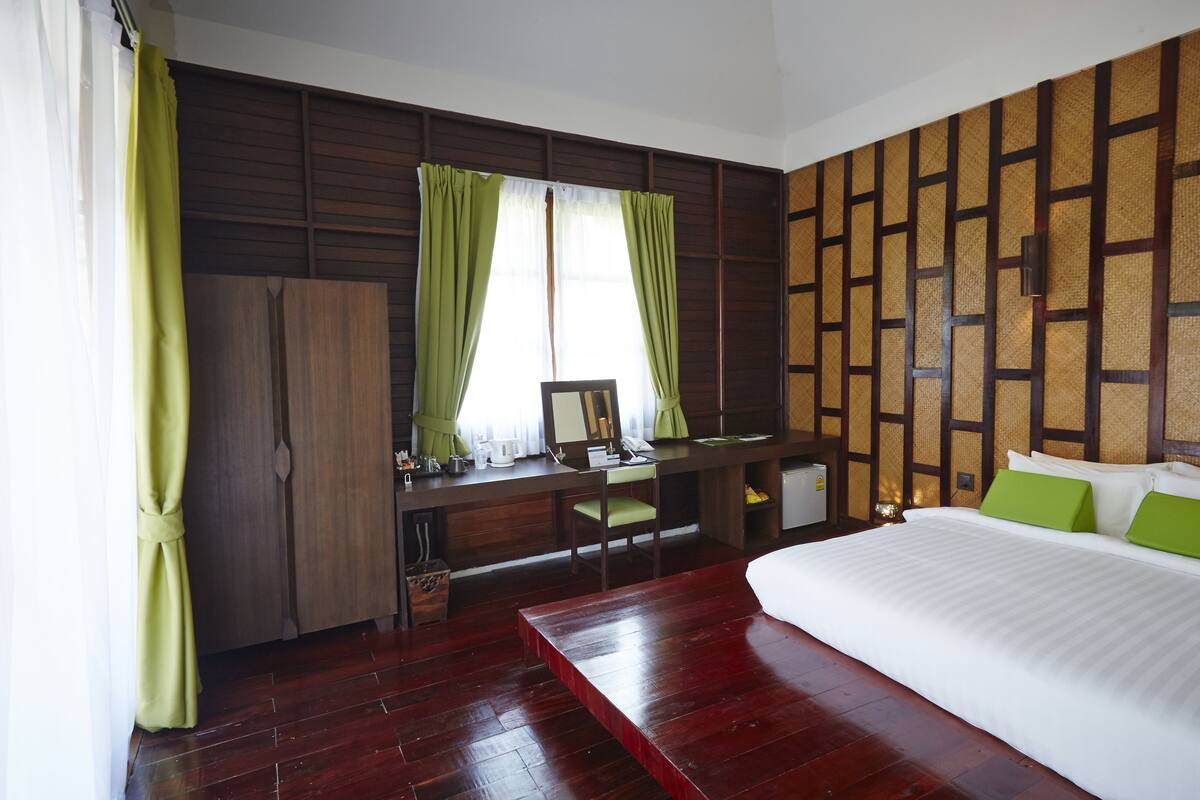 A hotel room with a wooden desk, a bed with green pillows, and a large window with curtains.