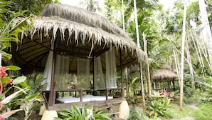 A thatched-roof hut with a wooden bench and a bed, surrounded by lush greenery and plants. - Wareerak Hot Spring & Wellness (Khlong Thom)