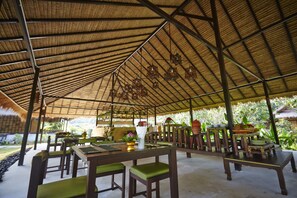 Un pavillon en plein air doté de tables et de chaises en bois, d’un toit de chaume et de plantes en pot.