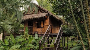 A wooden cabin with a thatched roof, surrounded by lush greenery and a wooden staircase leading to a balcony. - Wareerak Hot Spring & Wellness (Khlong Thom)