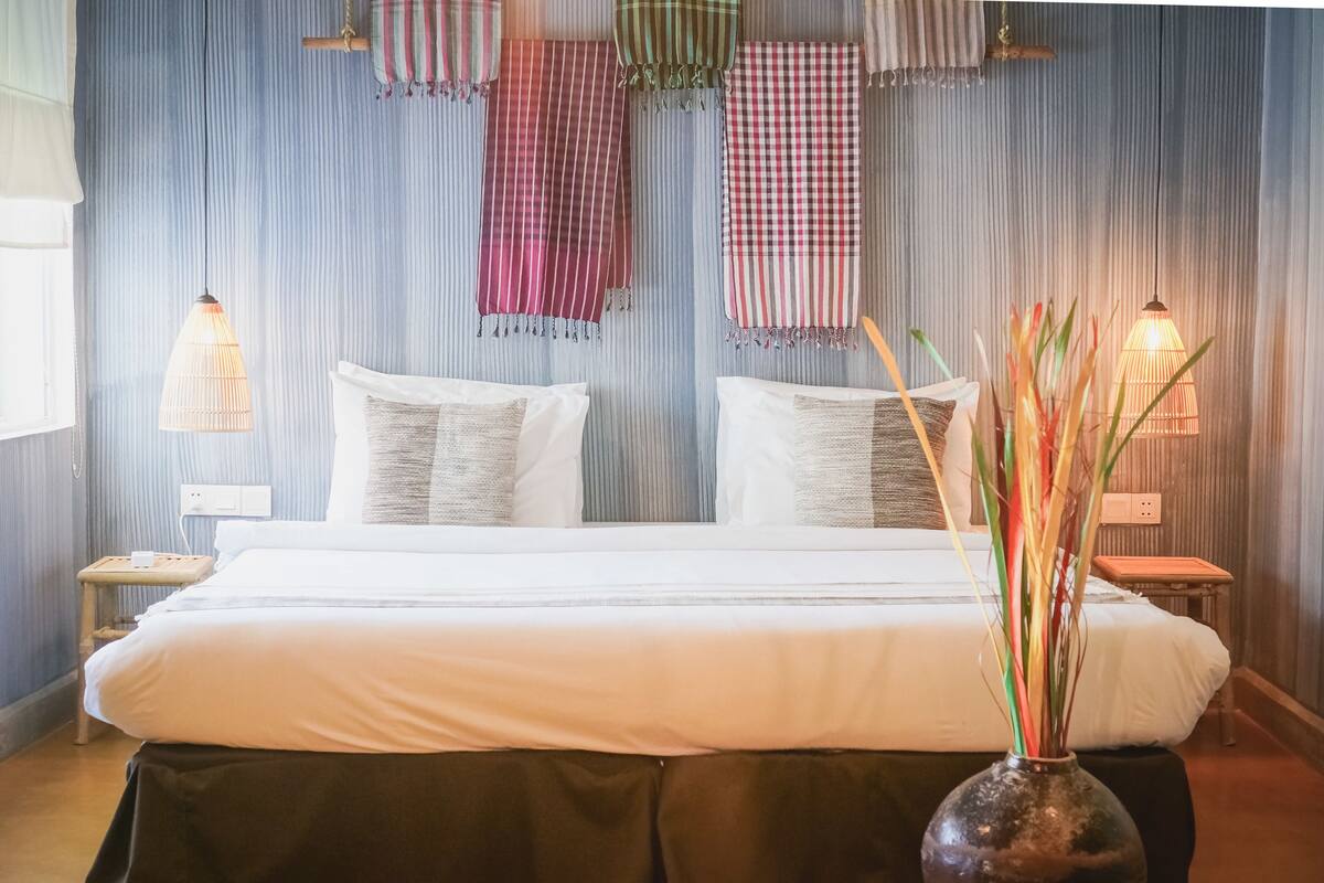 Een net opgemaakt bed met witte lakens, een vaas met hoge planten en gestreepte gordijnen aan de muur.