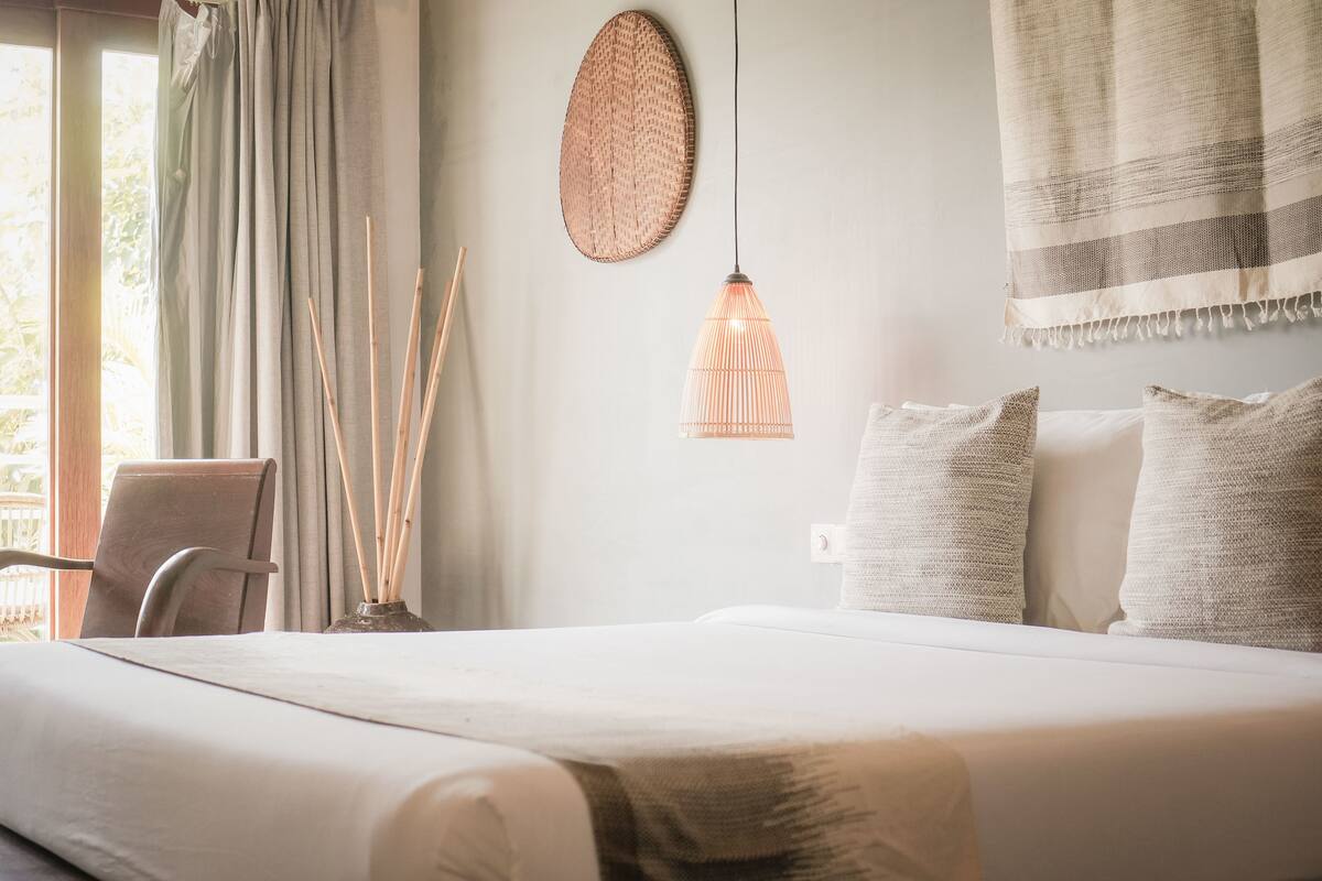 Een net opgemaakt bed met een witte deken, een hangend wandkunstwerk van geweven materiaal, een hanglamp en een stoel met kussen.