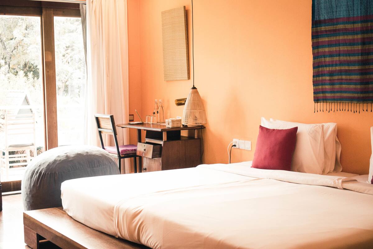 Een hotelkamer met een bed, een bureau en een stoel. Er is een raam met gordijnen, een wanddecoratie en een lamp.