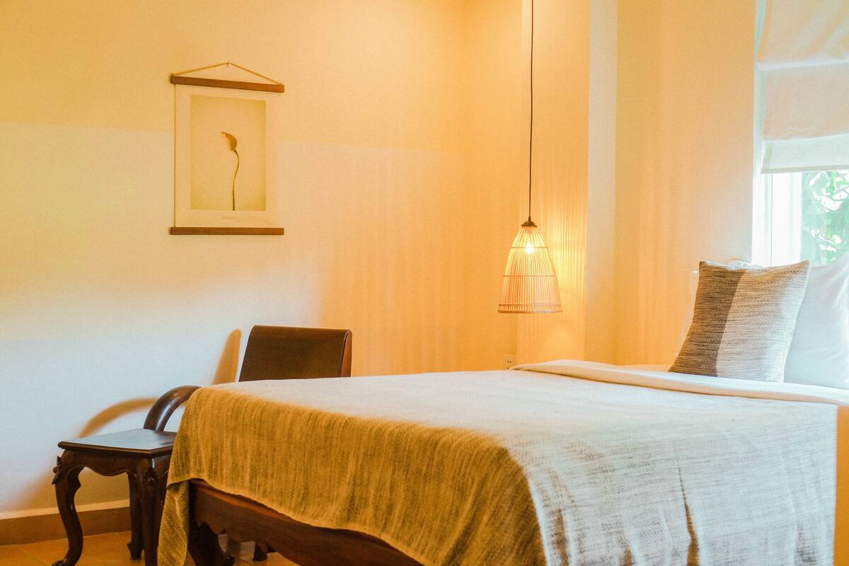 Een net opgemaakt bed met een wit-beige deken, een hangende lamp en een ingelijst foto aan de muur.