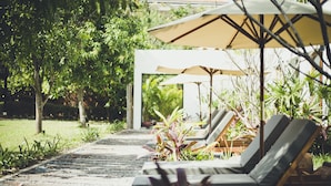 Un espace aménagé au bord de la piscine, avec des chaises longues et des parasols, entouré de verdure et bordé d’un sentier.