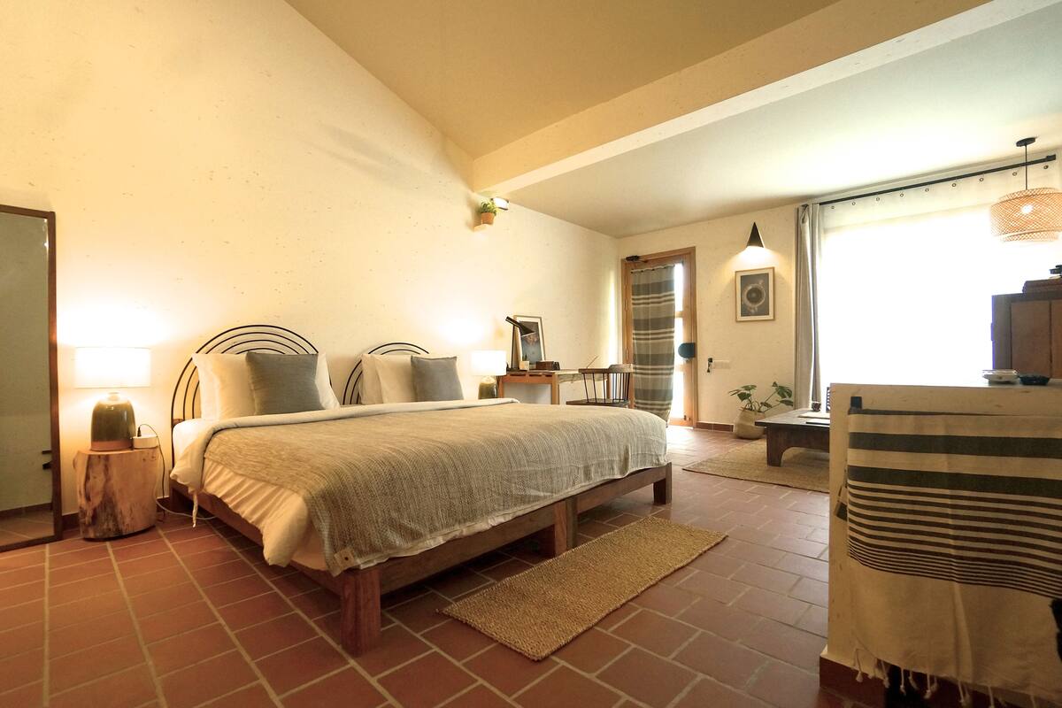 Een slaapkamer met een groot bed, nachtkastjes, een stoel, een kleine tafel en een raam met gordijnen.
