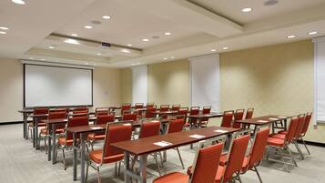 Un salón de conferencias con filas de mesas y sillas, una pantalla de proyector y una pizarra.