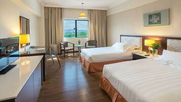 Quarto de hotel com duas camas, uma escrivaninha, uma cadeira, uma mesinha com um vaso e uma janela com cortinas.