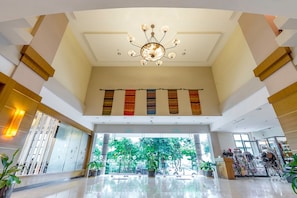 Um hall amplo com teto alto, um lustre central e vista para o exterior através de grandes janelas.