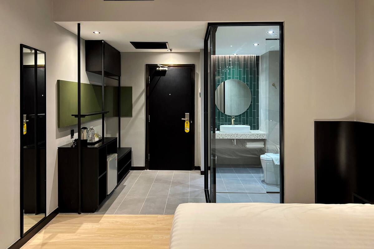 Une chambre d’hôtel moderne équipée d’un lit, d’un bureau, d’un miroir et d’une salle de bain visible à travers une porte en verre.