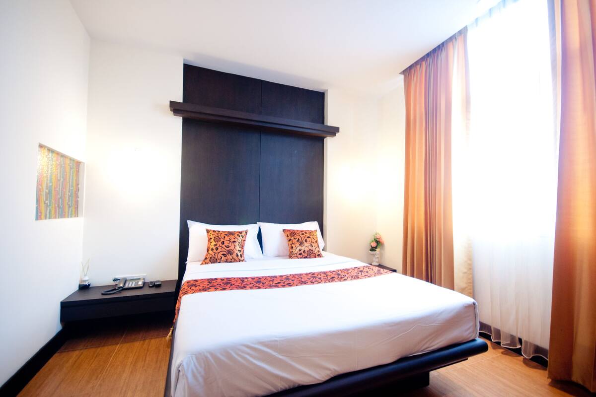 A hotel room with a large bed, a dark headboard, a bedside table with a phone and remote controls, and a window with curtains.