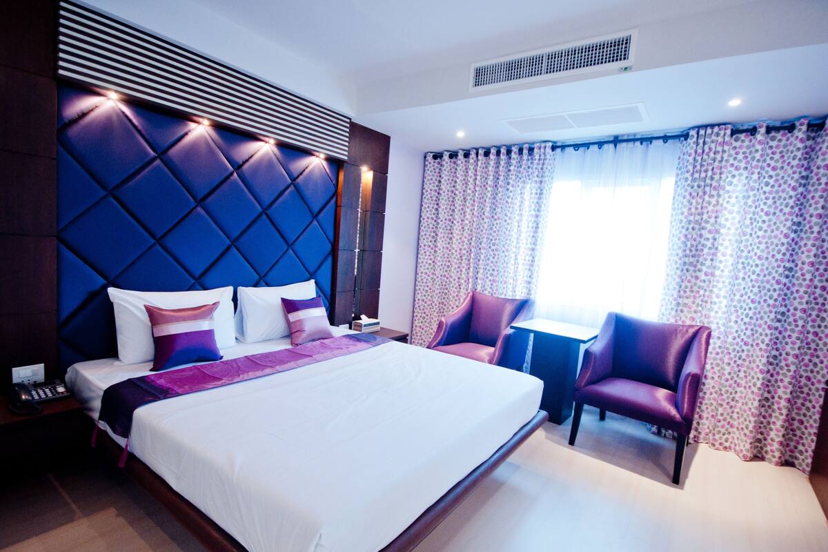 A modern hotel room with a large bed, a headboard with built-in lighting, a small table, two chairs, and a window with patterned curtains.