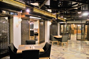 Ravintola, jossa on puupöydät ja -tuolit, koristeelliset sermit ja katosta roikkuvat valaisimet.