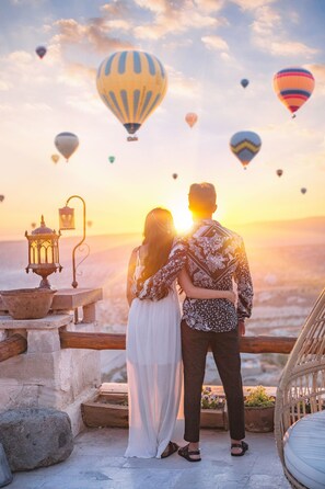 Un couple se tient sur un balcon, d’où il peut admirer un coucher de soleil et des montgolfières dans le ciel.