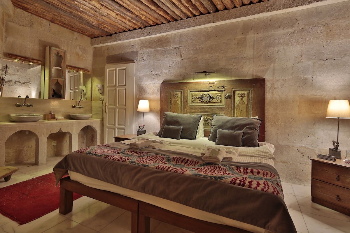 Une chambre à coucher avec un grand lit, une tête de lit en bois, deux lampes de chevet, une table de chevet, un miroir et un mur en pierre.