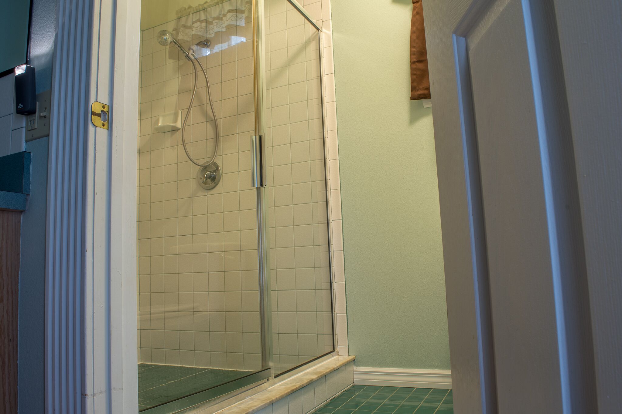 Ducha con mampara de vidrio, piso y paredes alicatadas, ducha visible y puerta que da a otra habitación.