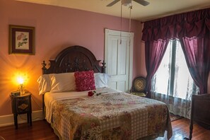 Un dormitorio con cama, mesita de noche con lámpara, un cuadro en la pared y una ventana con cortinas.
