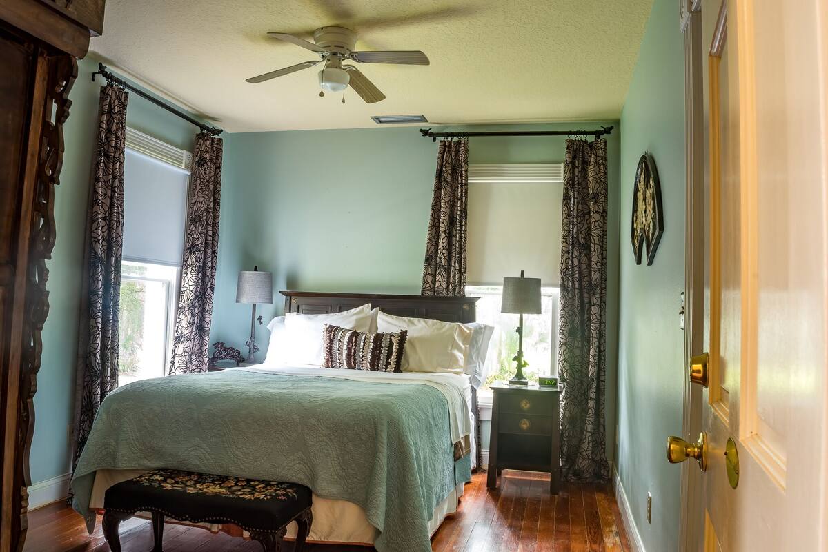 A bedroom with a bed, bedside tables, a bench, curtains, a ceiling fan, and a door.