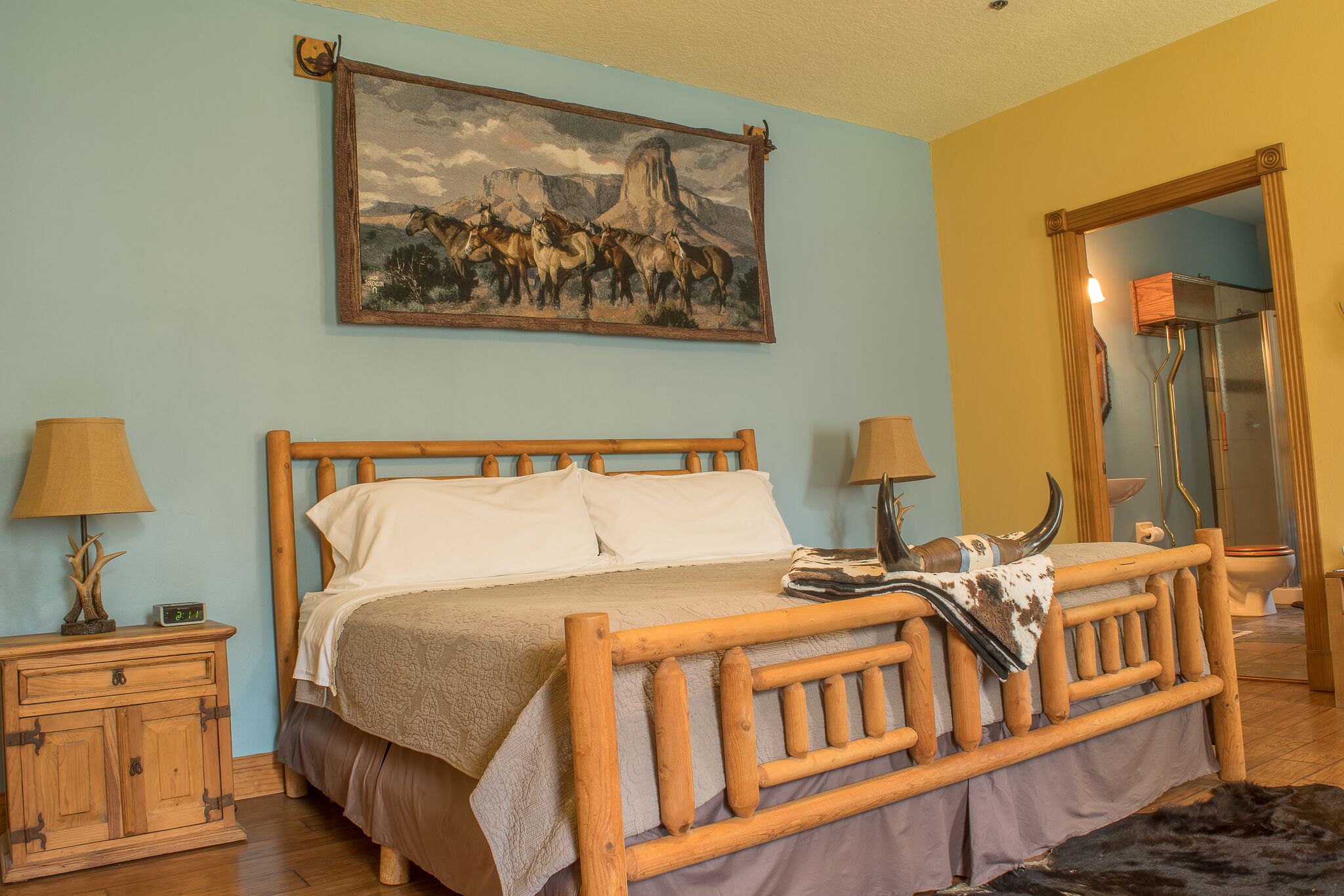 A wooden bed with a headboard, a nightstand with a lamp, and a painting of horses on the wall.