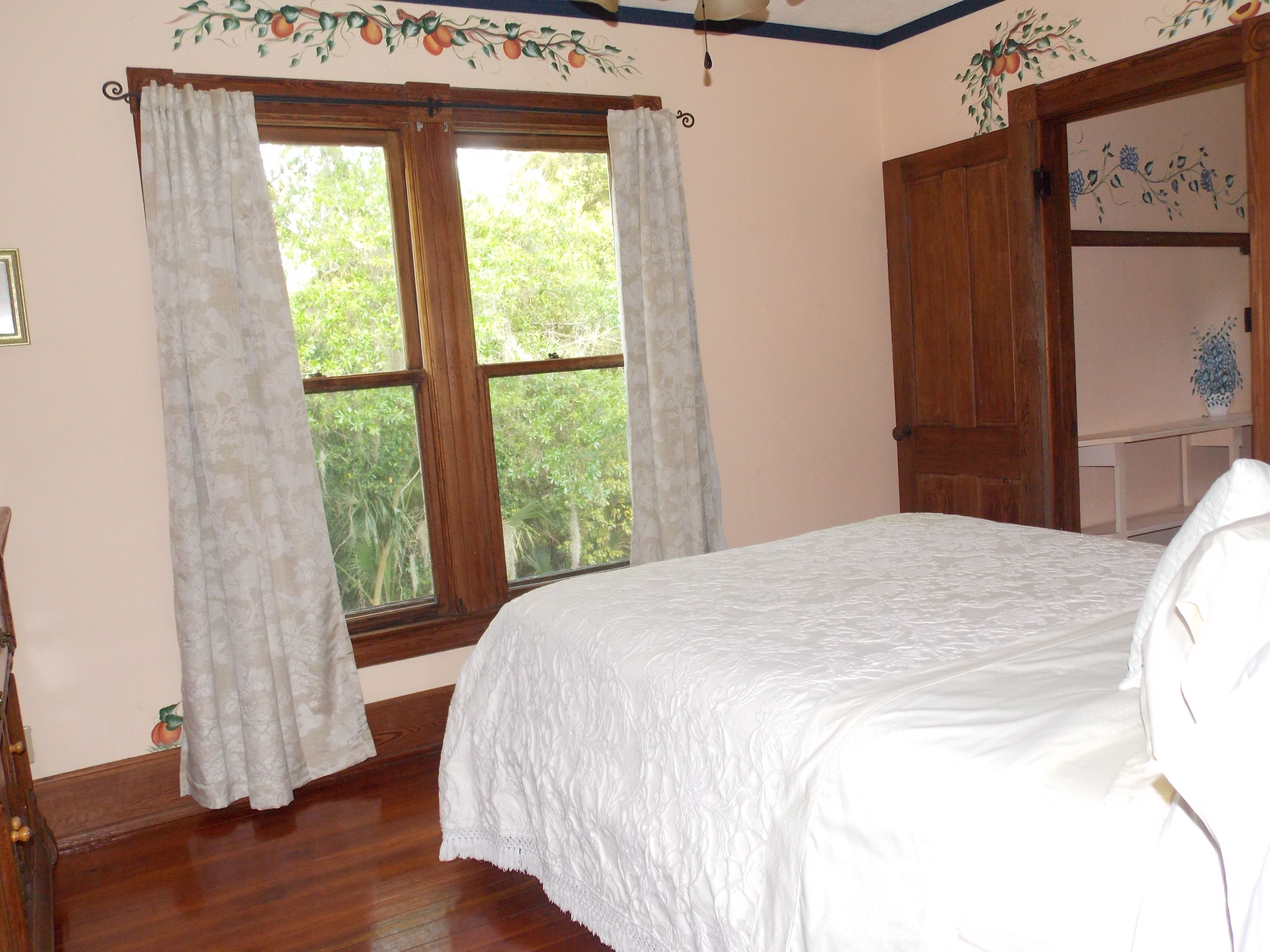 A bedroom with a bed, a window with curtains, a wooden wardrobe, and a wall with fruit and flower patterns.