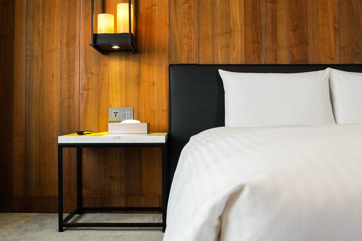Habitación de hotel con pared de madera, cama con ropa blanca, mesita de noche con lámpara y una pequeña mesa con un libro y un cuenco.