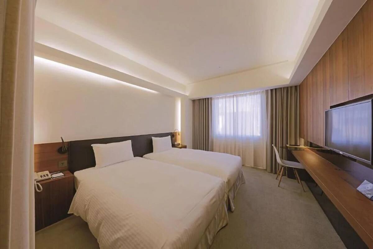 Habitación de hotel con dos camas, un escritorio, una silla, un televisor y una ventana con cortinas.