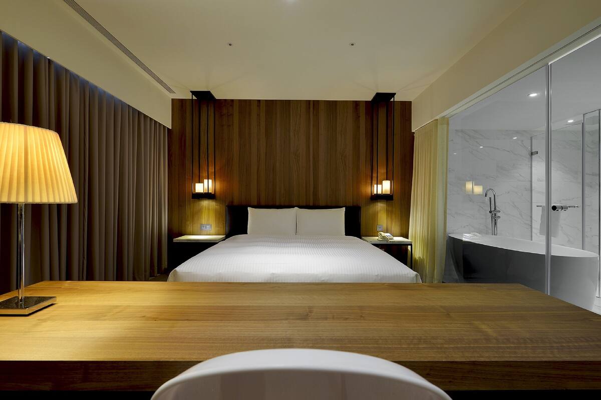 Una habitación de hotel moderna con una cama grande, paneles de madera, dos mesitas de noche con lámparas, un box de ducha de vidrio y un escritorio de madera.