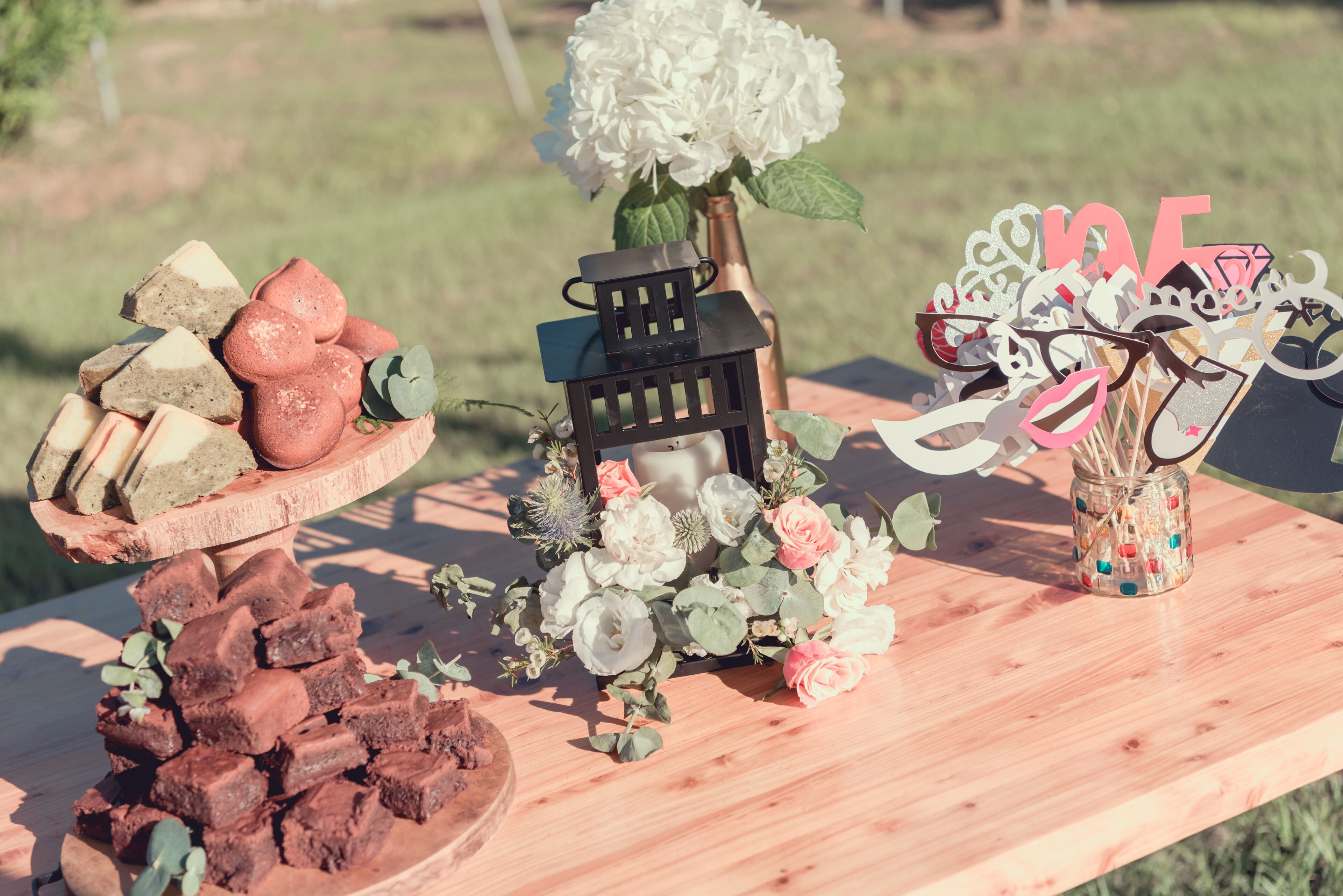 A wooden table with a variety of items including a cake stand with a cake, a decorative lantern, a vase with flowers, and a jar with a bow.