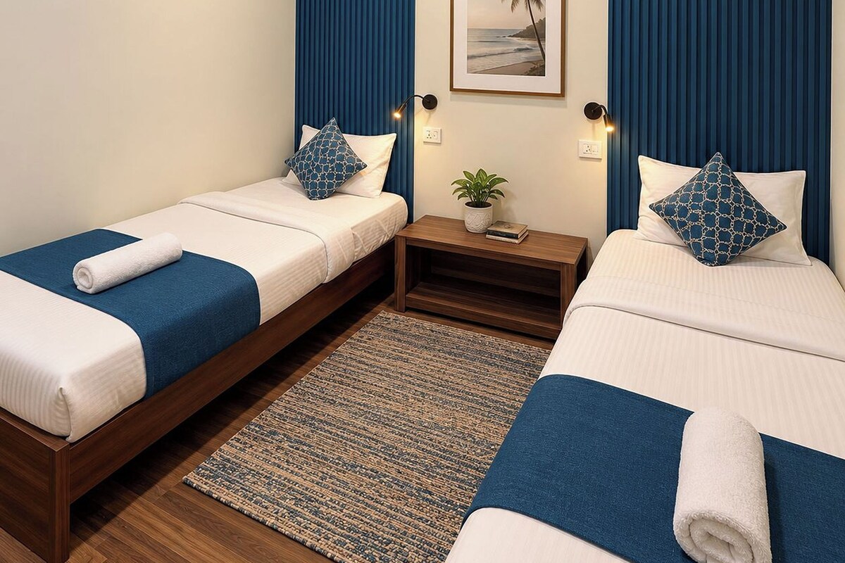 A hotel room with two single beds, a small bedside table with a plant, and a framed picture on the wall.