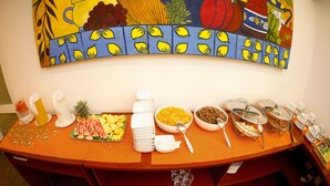 A buffet table with various food items and utensils, a fruit platter, and a colorful painting above. - CityRest Fort - Hostel (Colombo)