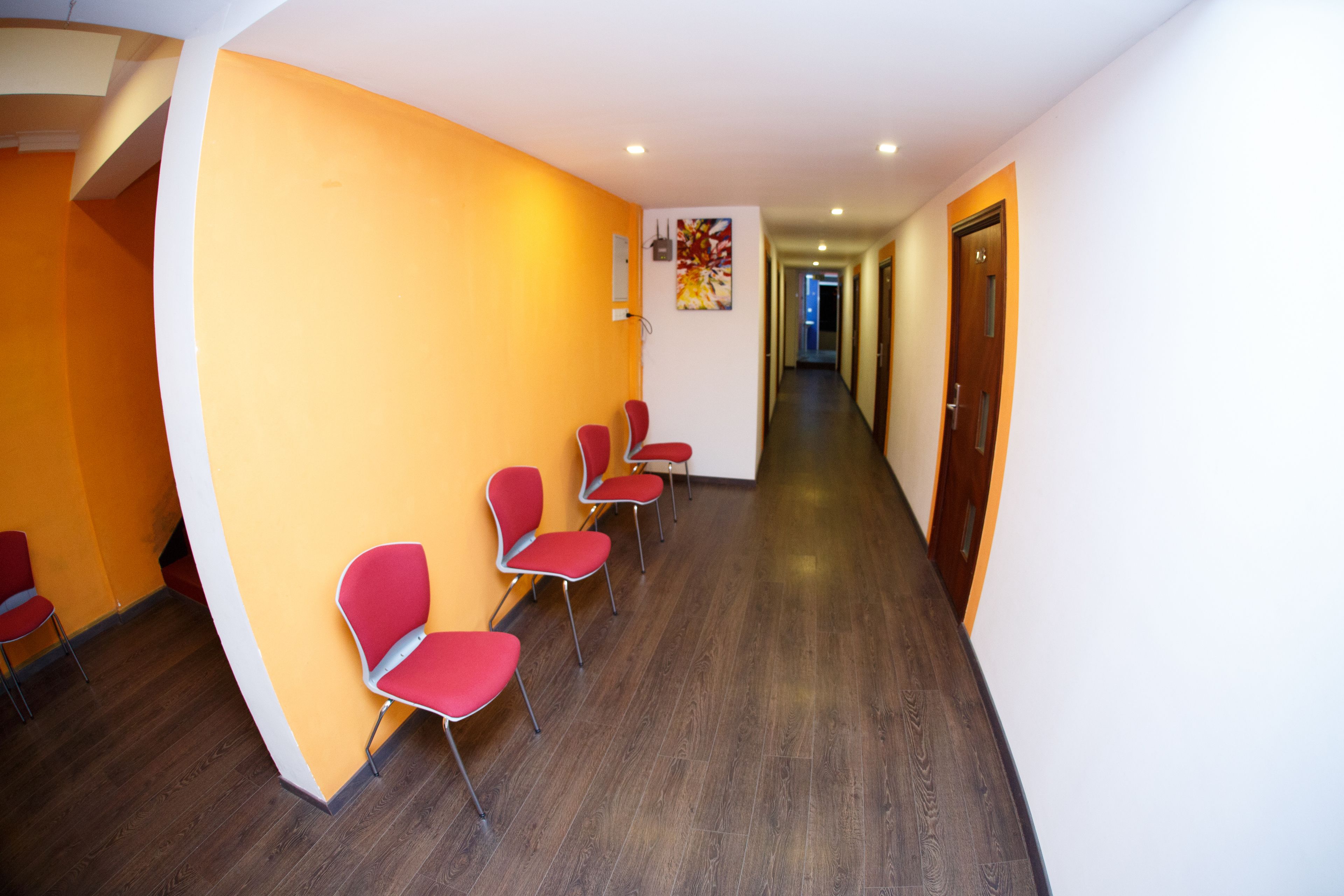 Un couloir où des chaises rouges sont alignées contre un mur jaune, et qui mène à une porte au fond.