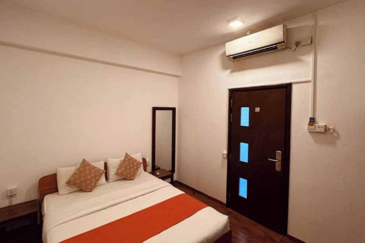 A hotel room with a bed, a mirror, a door with a light switch, and an air conditioning unit.