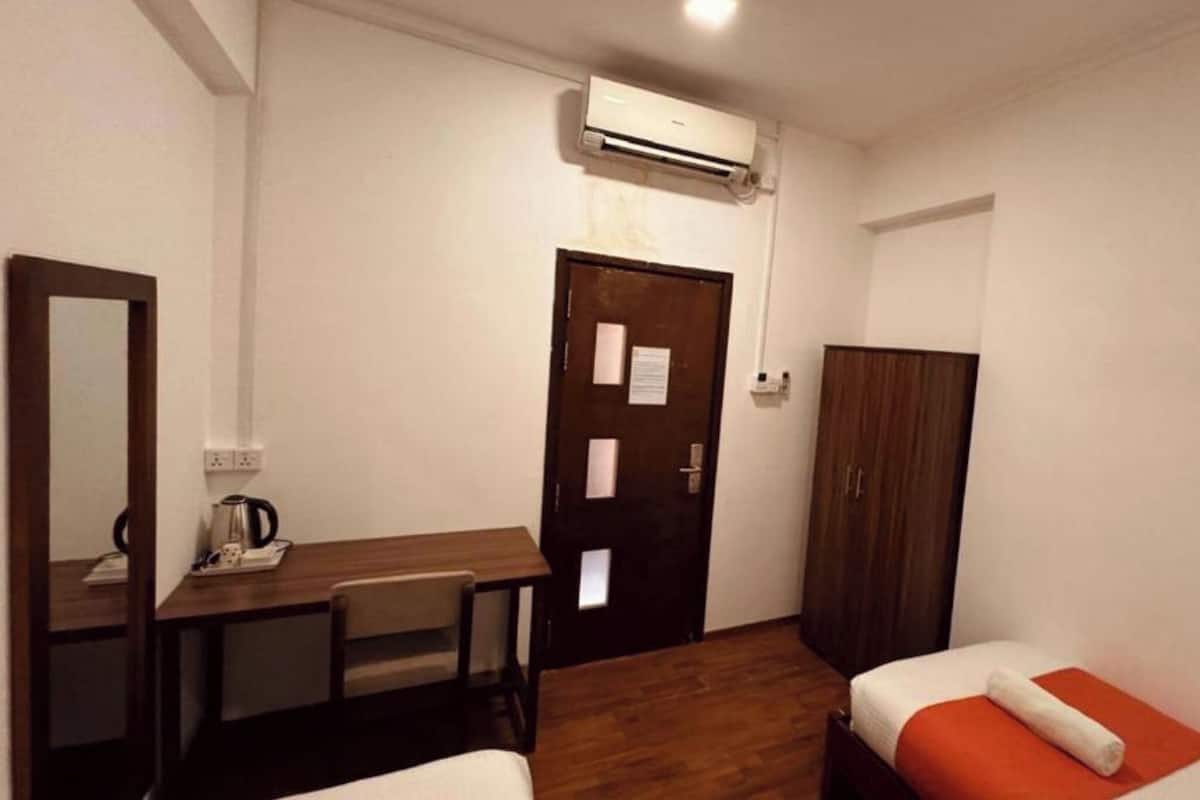 A compact hotel room with a wooden desk, two single beds, a wardrobe, and an air conditioning unit.