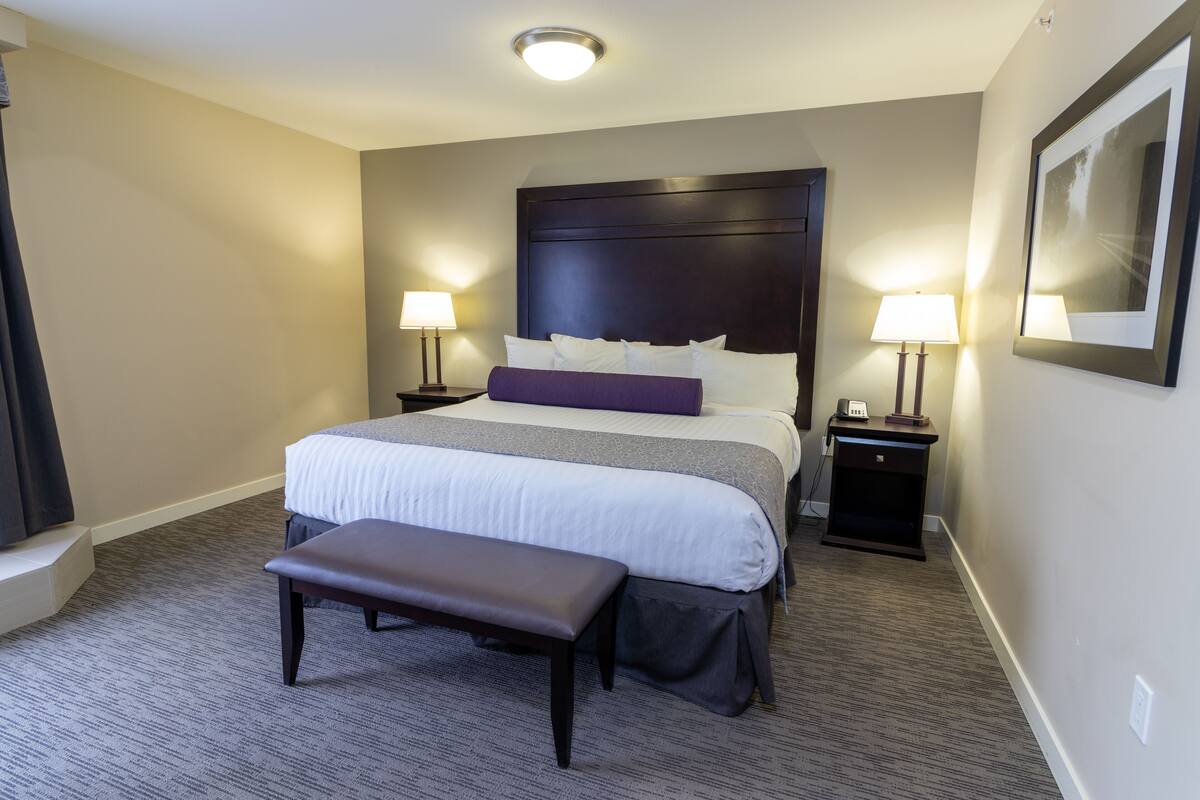 A hotel room with a large bed, two bedside tables with lamps, a bench, and a framed picture on the wall.