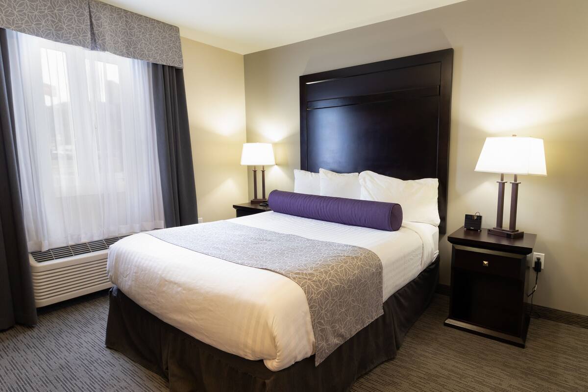 A hotel room with a large bed, two bedside tables with lamps, a window with curtains, and a wall-mounted heater.
