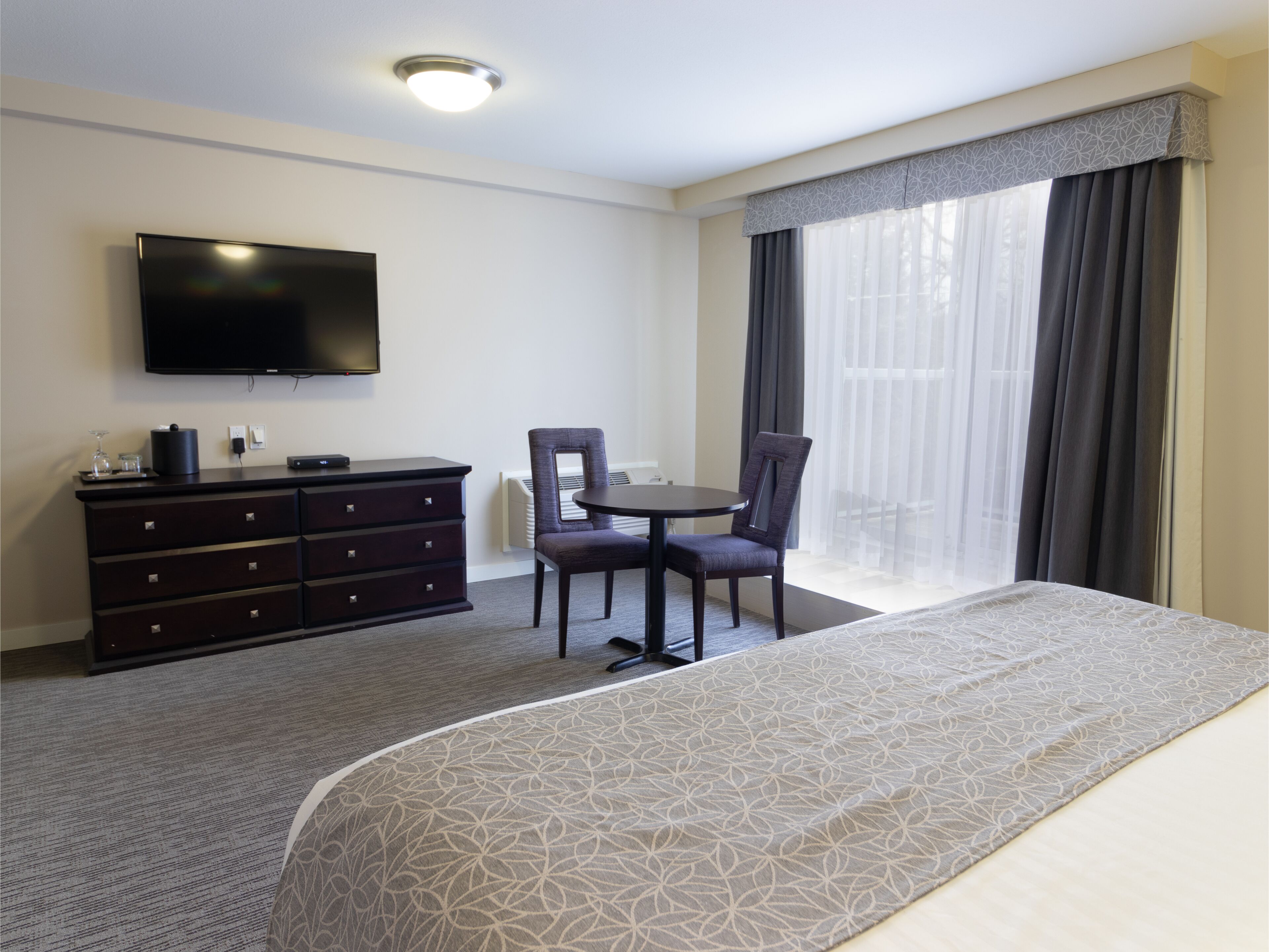 Habitación de hotel con cama, televisor montado en la pared, cómoda, silla y ventana con cortinas.