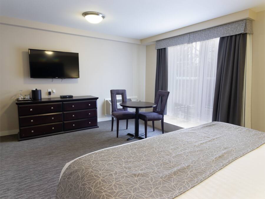Habitación de hotel con cama, televisor montado en la pared, cómoda, silla y ventana con cortinas.