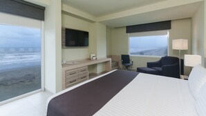 Habitación de hotel con una cama grande, un escritorio con computadora, una silla, un televisor y una ventana con vista a la playa.