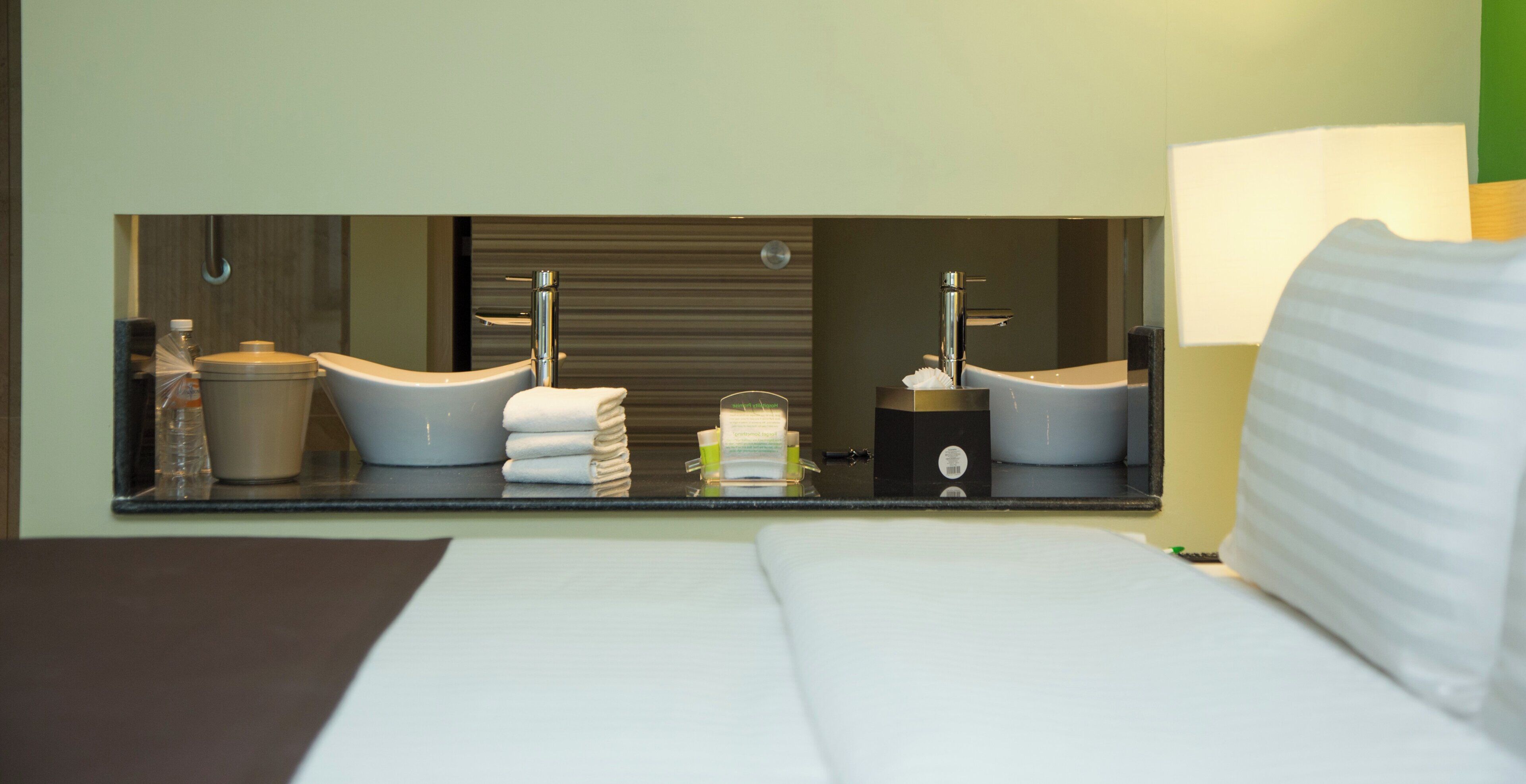 A hotel room with a bed, a large mirror reflecting a bathroom with two sinks, a toilet, and a towel dispenser.