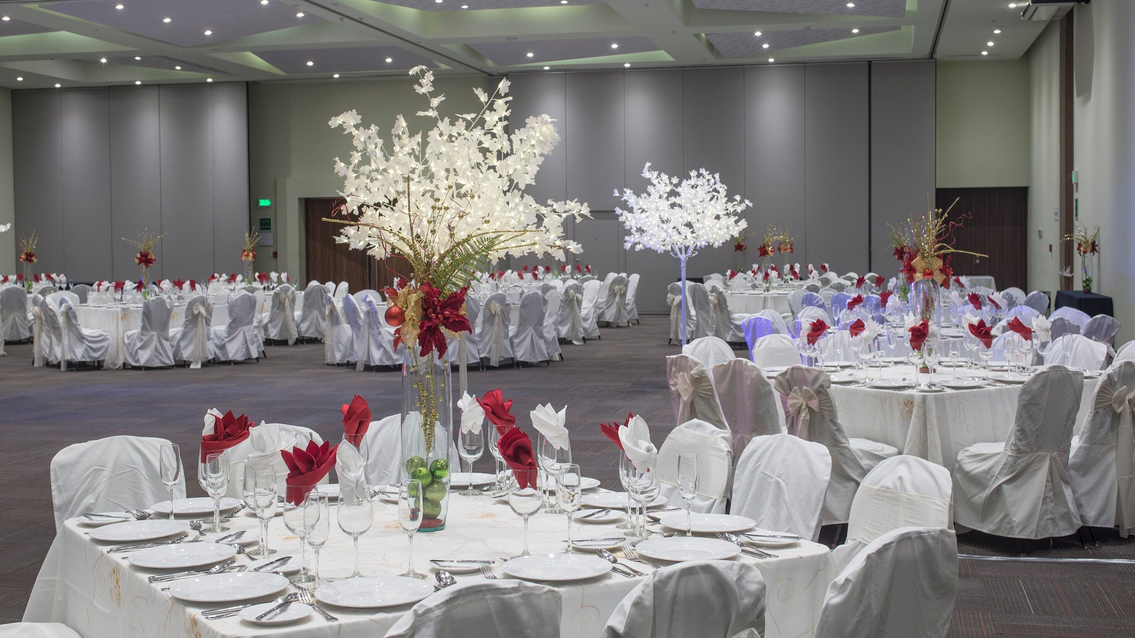 A banquet hall with round tables set for a formal event, featuring white tablecloths, glassware, and red napkins.