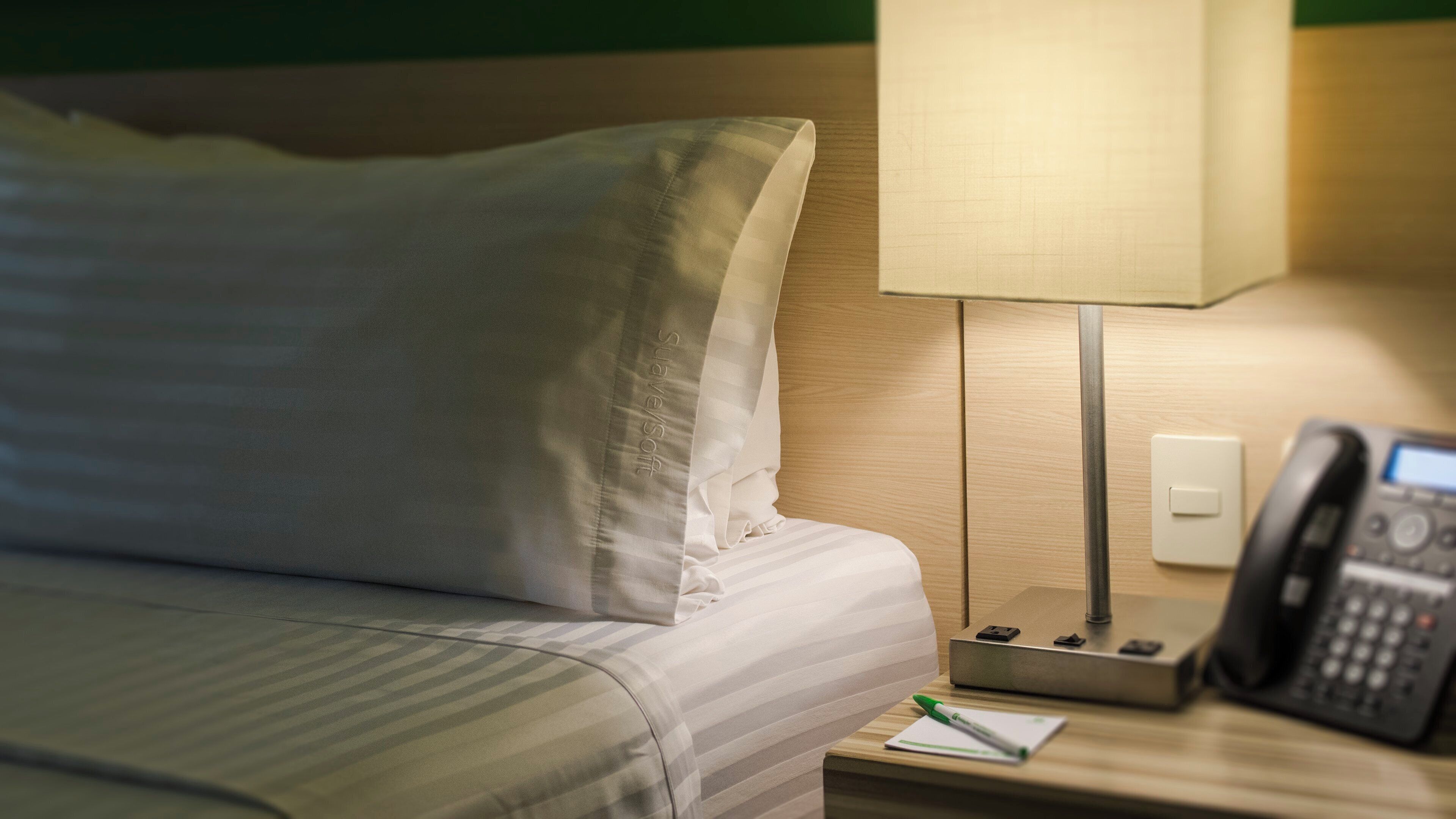 A hotel room with a bed, a bedside lamp, a telephone, and a notepad on the nightstand.