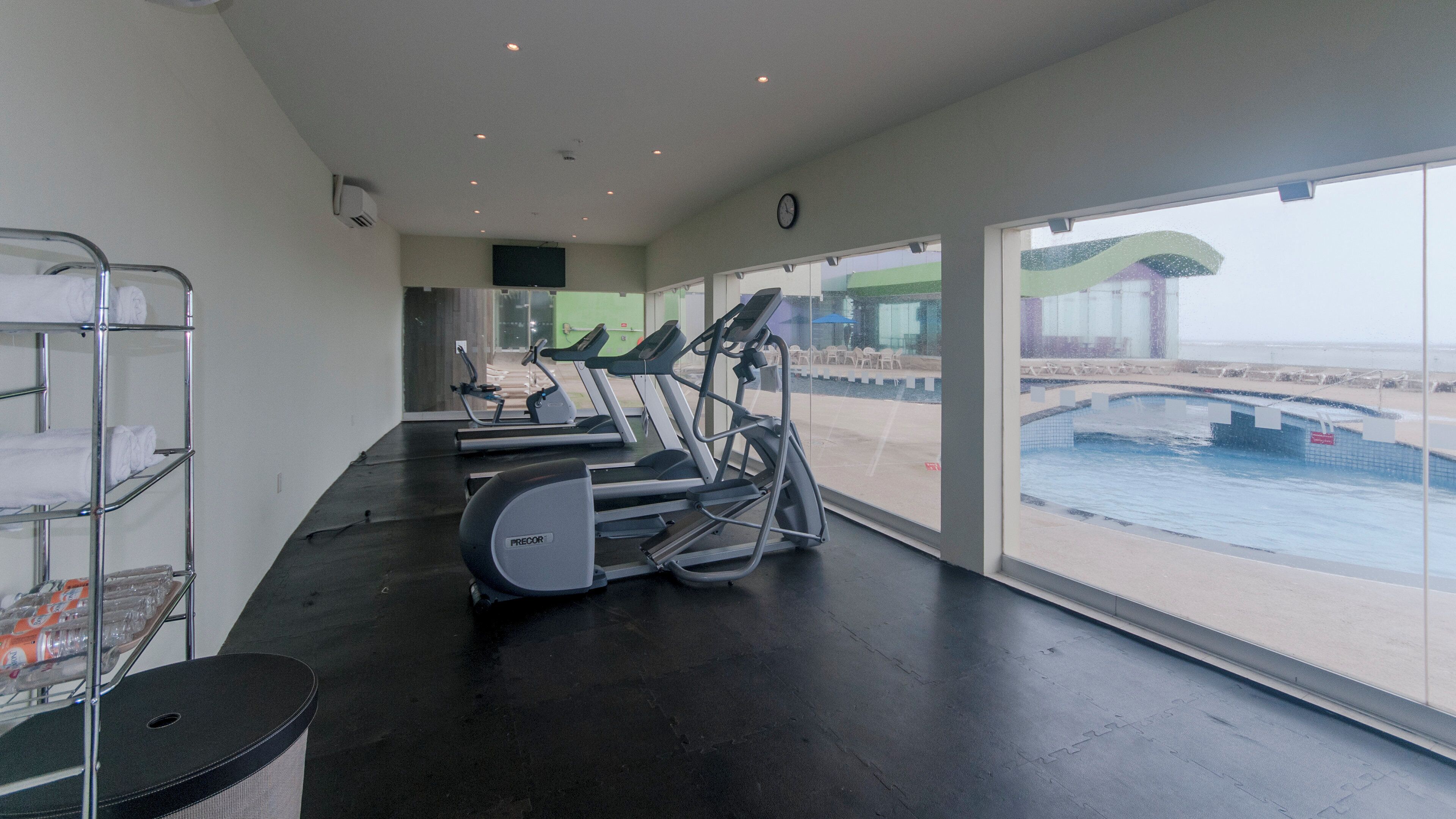 Un gimnasio con cintas de correr y equipo de ejercicio, ventanas amplias con vistas a una piscina y al mar.