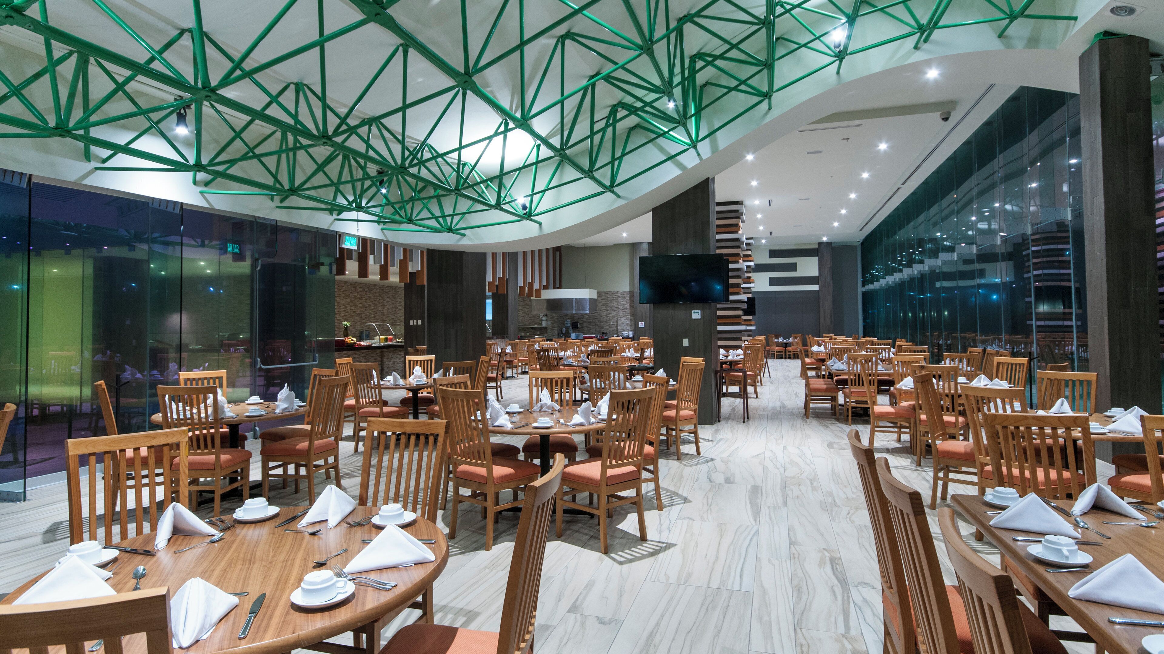 A spacious dining area with multiple tables and chairs, a modern ceiling design, and a large screen on the wall.
