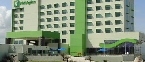 Un hotel Holiday Inn con diseño moderno, destacando un acento verde en la fachada, múltiples ventanas y un área exterior ajardinada con palmeras.