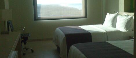 A hotel room with two beds, a desk, and a window with a view of the beach.