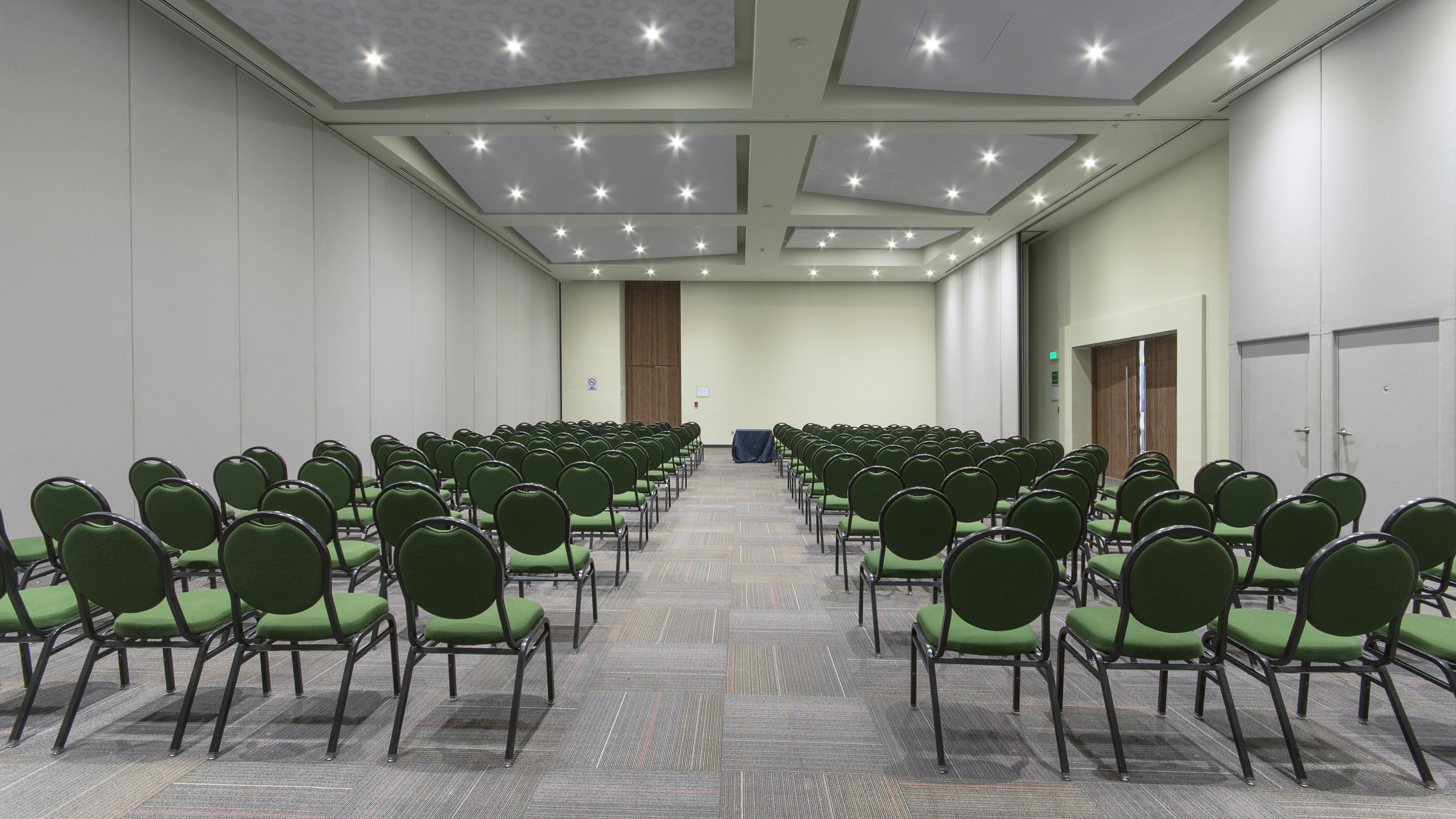 Une salle de conférence avec des rangées de chaises vertes, un sol recouvert de moquette et un plafond doté d’un éclairage encastré.