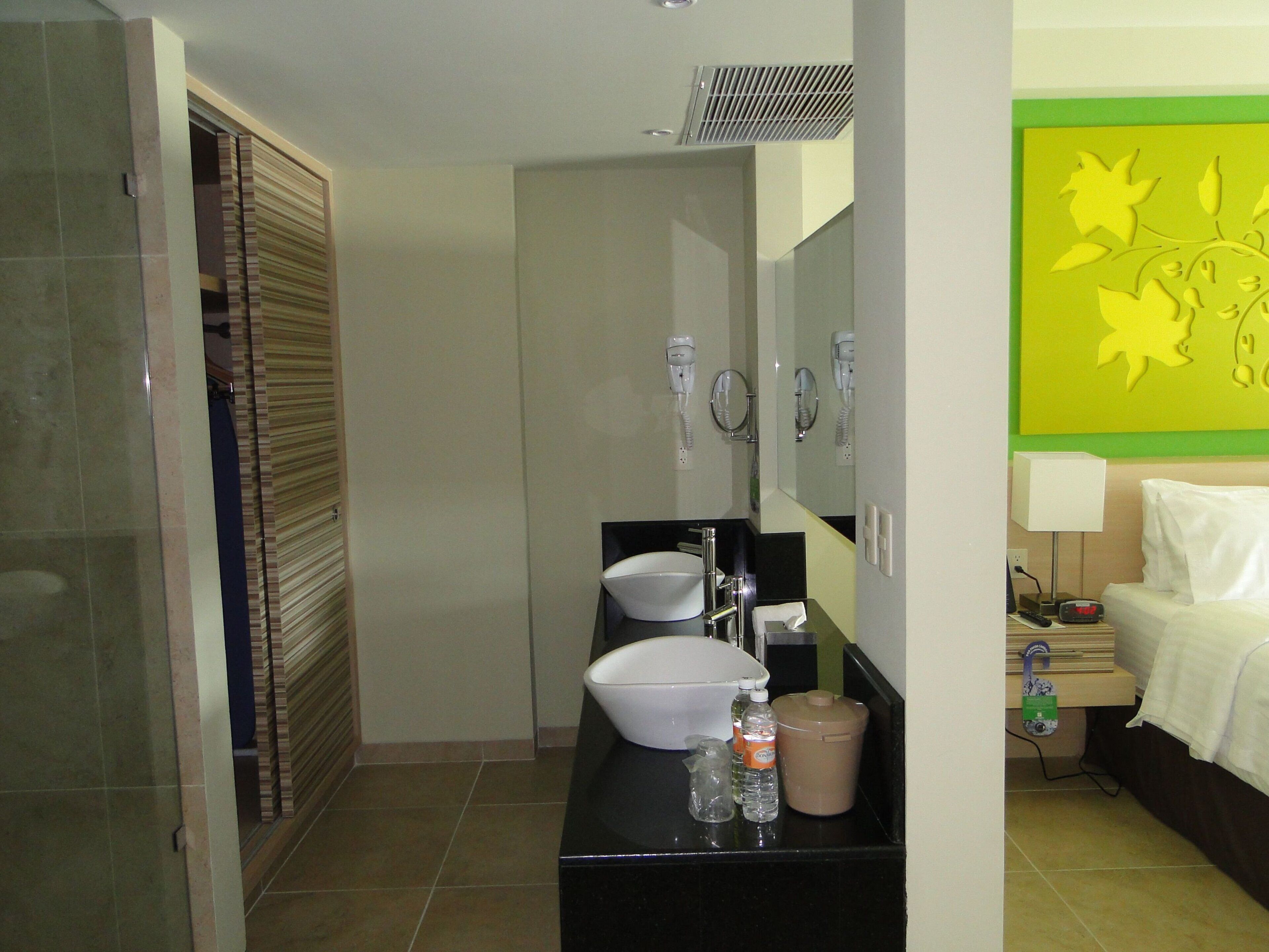 A hotel bathroom with a sink, mirror, hairdryer, and a bed with a nightstand.